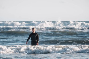 Dalgıç kıyafetli genç sahilde dalgaların arasında yürüyor.