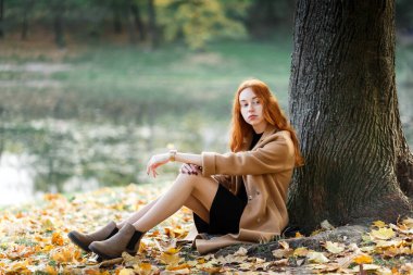 Şık kızıl saçlı kadın sonbaharda güneşli bir parkta yürüyor.