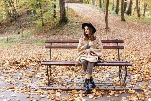 秋には黒の帽子を被った茶色の髪の女性とベンチのポンチョ — ストック写真