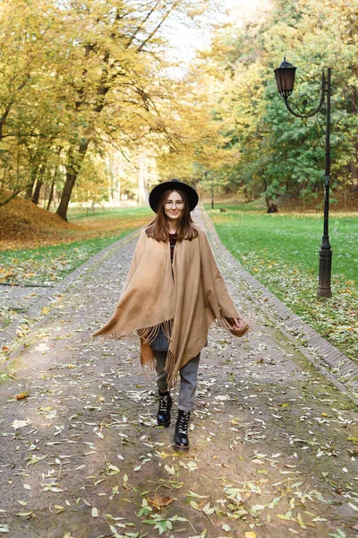 黒の帽子をかぶった茶色の髪の女性と秋の公園を歩くポンチョ — ストック写真