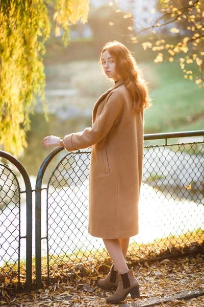 コート姿の赤髪の女性が秋の晴れた公園を歩く — ストック写真