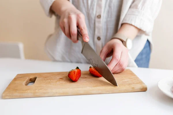 female cook cuts red fresh strawberries with knife in kitchen