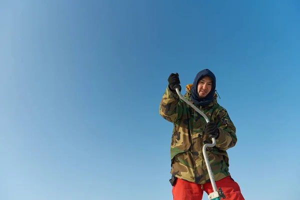 A warmly dressed Asian guy drills a hole in the ice on winter fishing