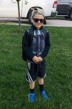 Gülümseyen şapkalı ve güneş gözlüklü çocuk, okul macerasına hazır.