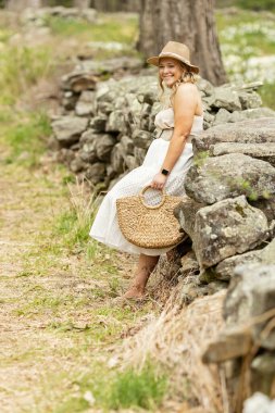 Gülümseyen kadın, taş duvara yaslanmış, şapka takıyor.