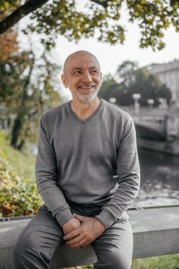 Yaşlı bir adam nehir kenarındaki bankta oturuyor, gülümsüyor, günün tadını çıkarıyor.