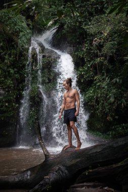 Tişörtsüz bir adam, ağaç kütüğünde yürüyor, Tayland 'da bir şelalede.