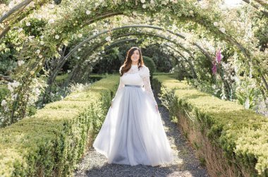 Bahçede çiçeklerin altında mavi elbiseli neşeli kadınlar yürüyor.