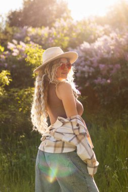 Şapkalı ve güneş gözlüklü seksi kıvırcık sarışın çiçek açan ağaçların yanında poz veriyor.