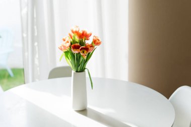 Güneş ışığında yuvarlak bir masada, beyaz vazoda turuncu laleler..