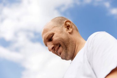 Beyaz tişörtlü adam gülümsüyor ve mavi gökyüzüne bakıyor.