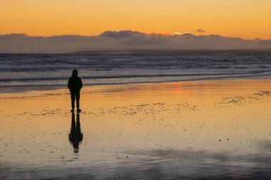 Gün batımında sahilde duran kişi ıslak kumda yansıyor.
