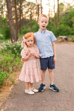 Kızıl saçlı kız ve erkek kardeş çiçeklerin yanında dururlar.