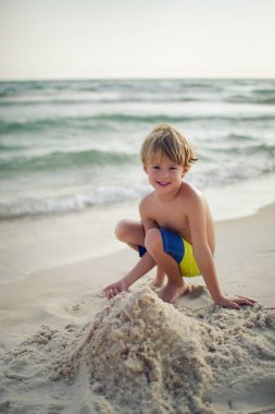 Güneşli bir sahilde kumdan kalesi olan gülümseyen çocuk.