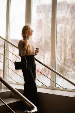 Gözlüklü genç bir kadın merdivenlerde kahve içiyor.