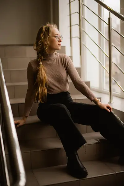 stock image A young woman in glasses sits on the stairs and looks out the window