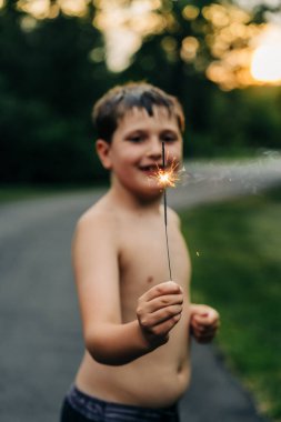 Close-up of a boy holding a lit sparkler, smiling at sunset clipart