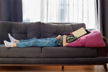 Çocuk kanepede uzanıyor, yüzünde bir kitapla, uyuyor.