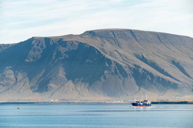Büyük bir kayalık dağın yanında yelken açan balıkçı teknesi.