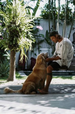 Adam bahçede salıncakta sallanırken bir golden retriever tutuyor..