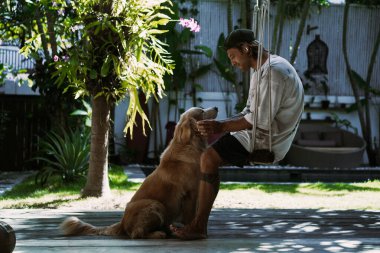 Adam bahçede salıncakta sallanırken bir golden retriever tutuyor..