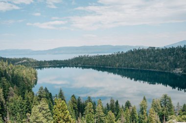 Uzakta dağları olan bir gölün üzerinde yansıyan ağaçların görüntüsü.