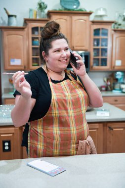 Woman talking on phone while holding a pen in a kitchen. clipart