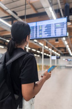 Genç bir kadın telefonu kontrol ediyor ve havaalanı ekranlarına bakıyor.