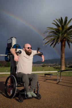 Adult male wheelchair user exercising outdoors under rainbow clipart