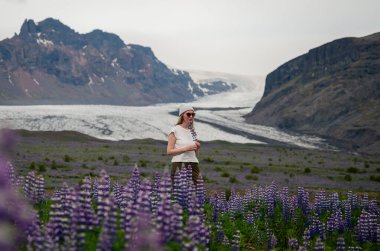 young woman stands in a lupine field in front of a glacier in Iceland clipart