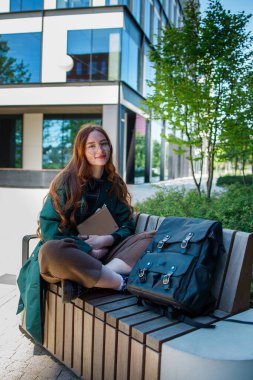 Sırt çantası ve kitaplarıyla bankta oturan kızıl öğrenci kız.