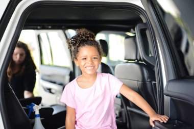 Smiling child inside a car with an adult cleaning in the backgro clipart