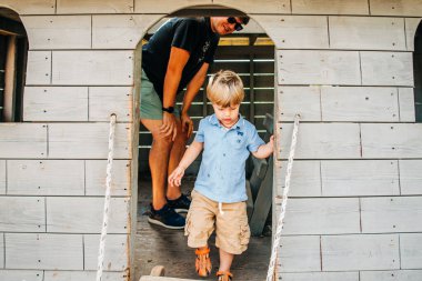 Young boy exiting a wooden playhouse with adult supervision clipart