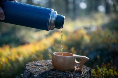 human hand pours tea from a thermos into a wooden mug in the forest clipart