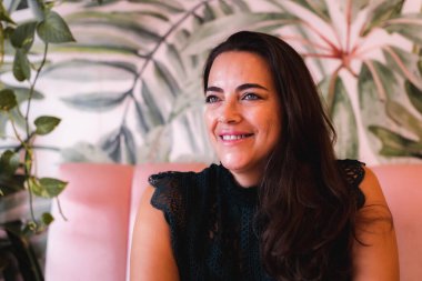 40 'larında, pembe bir koltukta oturan gülümseyen bir kadın.