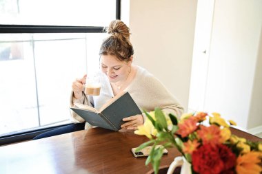 Pencerenin kenarında kitap okurken kahve içen genç bir kadın.