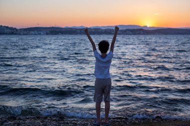 Çocuk gün batımında deniz manzarasında muzaffer bir şekilde kollarını kaldırıyor.