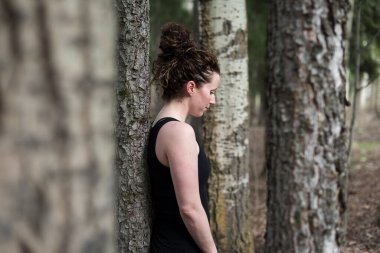 Düşünceli bakışları olan genç bir kadın ormanda ağaca yaslanıyor.
