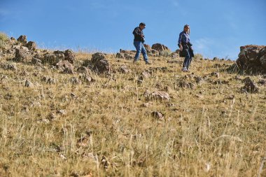 Açık mavi bir gökyüzünün altında kayalık bir yamaçta yürüyüş yapan iki kişi..