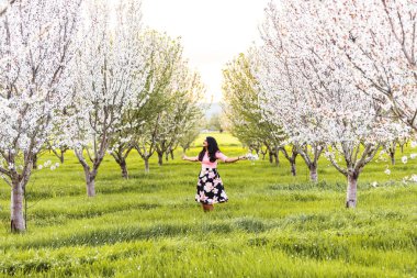 Çiçekli bir badem bahçesinde kolları açık duran bir kadın.