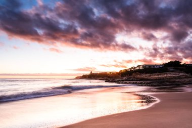 Dalgalar ve dramatik bulutlarla kumsalda huzurlu bir gün batımı