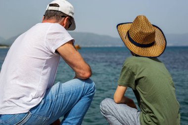 Deniz kenarında oturan bir adam ve bir çocuğun arka görüntüsü, ikisi de şapka takıyor.