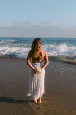 Bronz tenli kadın arkasında deniz kabuğu tutarken okyanusa bakıyor.
