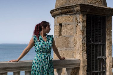 Kızıl saçlı kadın arka planda deniz olan korkuluklara yaslanıyor.