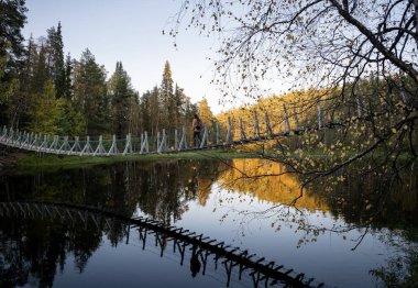 man in a yellow jacket crossing a suspension bridge in Finland clipart