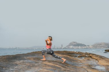 Genç bir kadın sahilde, dışarıda kayalarla yoga yapıyor.
