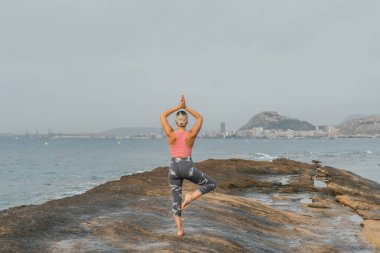 Genç bir kadın sahilde, dışarıda kayalarla yoga yapıyor.