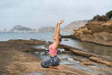 Genç bir kadın sahilde, dışarıda kayalarla yoga yapıyor.