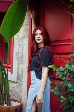 Kırmızı kapıya dayanmış, uzun kızıl saçlı genç bir kadın.
