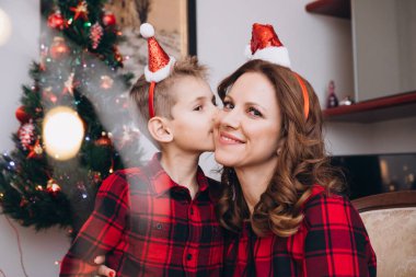 Ağacın yanında Noel kıyafetleri içinde anne ve oğluna sarılıp öpüyor.